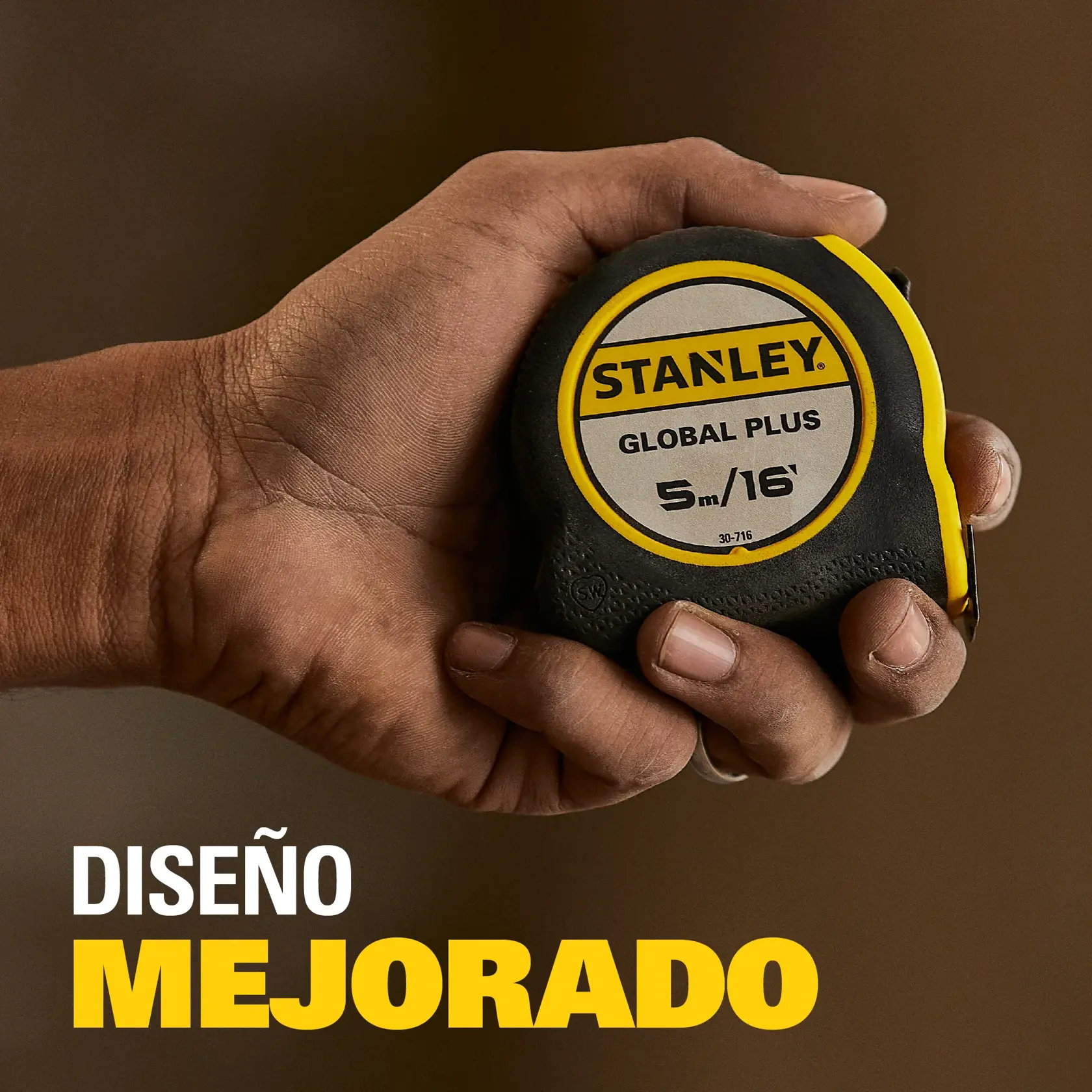 Primer plano de una mano sosteniendo un flexómetro Stanley Global Plus de 5 metros o 16 pies. El fondo está desenfocado. En la parte inferior izquierda hay texto en español que dice 'DISEÑO MEJORADO'.